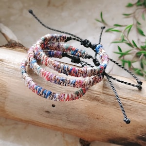 May include: Three colorful braided bracelets with black cords and adjustable knots. The bracelets are made of woven fabric with metallic threads and are stacked on a wooden surface.