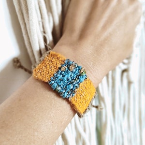 Puede incluir: Un brazalete tejido con un diseño amarillo y azul. El brazalete está hecho con una variedad de hilos y tiene una superficie texturizada.