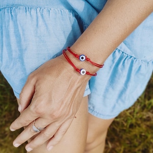 Könnte beinhalten: Zwei rote Schnurarmbänder, jedes mit einem blau-weißen Evil-Eye-Anhänger. Die Armbänder werden an einem Handgelenk getragen, wobei ein silberner Ring an einem Finger sichtbar ist. Der Hintergrund ist ein hellblaues Kleid und grünes Gras.