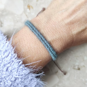 Puede incluir: Primer plano de una pulsera azul-verdosa hecha de un material tejido y texturizado. La pulsera está envuelta alrededor de una muñeca, con un cierre de cuerda marrón. La pulsera es el punto focal, con una manga borrosa de color púrpura claro en el fondo.
