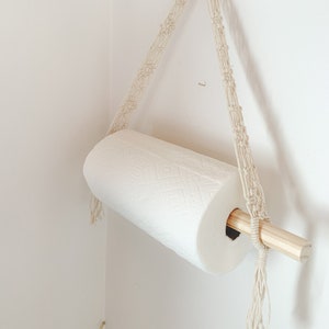 以下が含まれることがあります： マクラメの壁掛けホルダーに木製の棒が取り付けられ、白いペーパータオルのロールがぶら下がっています。ホルダーは天然の綿ロープで作られており、上部に木製のリングが付いています。
