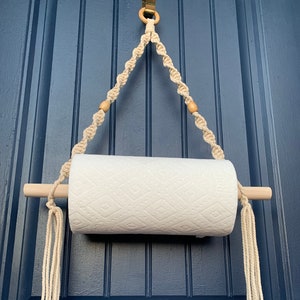 May include: A white paper towel roll hanging on a macrame paper towel holder with wooden dowels and natural colored tassels. The holder is hanging on a blue door.