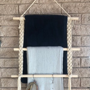 May include: A wooden wall-mounted shelf with three tiers for hanging items. The shelf is made of natural wood and has a macrame design. Two blankets are hanging on the shelf, one is black and the other is light gray. A belt and a decorative piece are hanging on the bottom tier.