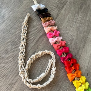 Op de afbeelding: Een lang, beige macramékoord met een geknoopt uiteinde. Een rij kleurrijke haarstrikken is aan het koord bevestigd, beginnend met wit en overgaand naar rood, oranje, geel, groen en blauw.