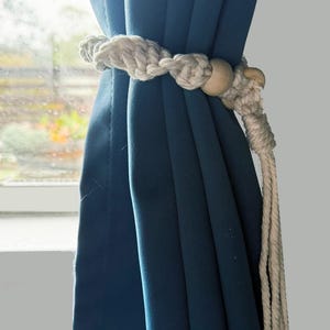 May include: A teal curtain is held by a white rope tieback with wooden beads. The rope is knotted and has a tassel. The curtain is in front of a window.