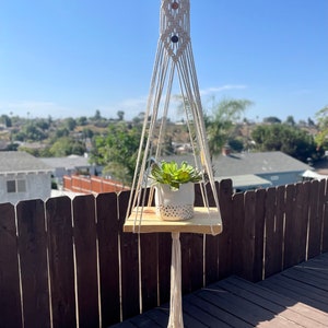May include: A macrame plant hanger with a wooden shelf and a potted succulent. The hanger is made of white cotton cord and has a beaded detail. The shelf is made of natural wood and is about 25 cm wide.