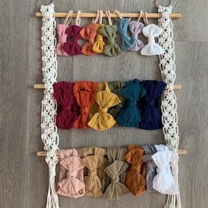 Op de afbeelding: Een houten wandorganizer met macramé hangende koorden bevat een verzameling kleurrijke stoffen hoofdbanden met strikken. De hoofdbanden zijn in rijen gerangschikt en zijn in tinten rood, oranje, geel, groen, blauw en wit.