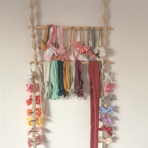 Op de afbeelding: Een houten stok met twee rijen kleurrijke hoofdbanden en haarbandjes die eraan hangen. De stok hangt aan twee macramé koorden met gouden ringen aan de bovenkant. Aan een van de koorden is een rij kleurrijke haarclips bevestigd.