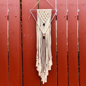 Pode incluir: Uma tapeçaria de macramé branca com contas de madeira e verdes, pendurada em uma moldura triangular de metal contra um fundo de madeira vermelha.