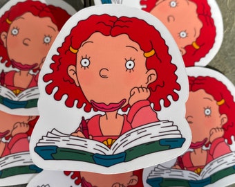 May include: A set of five stickers featuring a cartoon girl with red curly hair, wearing a pink shirt and holding a book. She is writing in the book with a red pen. The stickers are on a gray tiled surface.