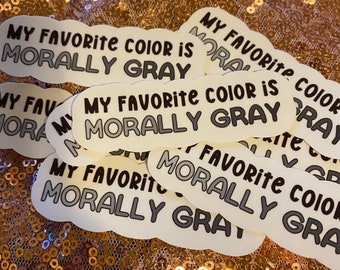 May include: Multiple white stickers with the text "My Favorite Color is Morally Gray" in black lettering. The stickers are in a cloud shape and are on a gold sparkly background.