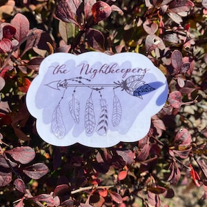 May include: A white sticker with a purple cloud shape and the text "the Nightkeepers" in black script. The sticker features a black and white illustration of an arrow with feathers.