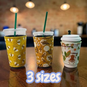 May include: Three beverage cups with fabric sleeves in different patterns and sizes. The left cup has a yellow bee print, the middle cup has a mustard floral print, and the right cup has a white monkey print. Each cup features a lid and straw.