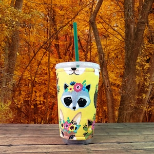 Puede incluir: Un protector de taza reutilizable amarillo con un dise&ntilde;o floral que presenta un mapache, un zorro y un conejo. El protector est&aacute; sobre una mesa de madera con un fondo borroso de follaje oto&ntilde;al.