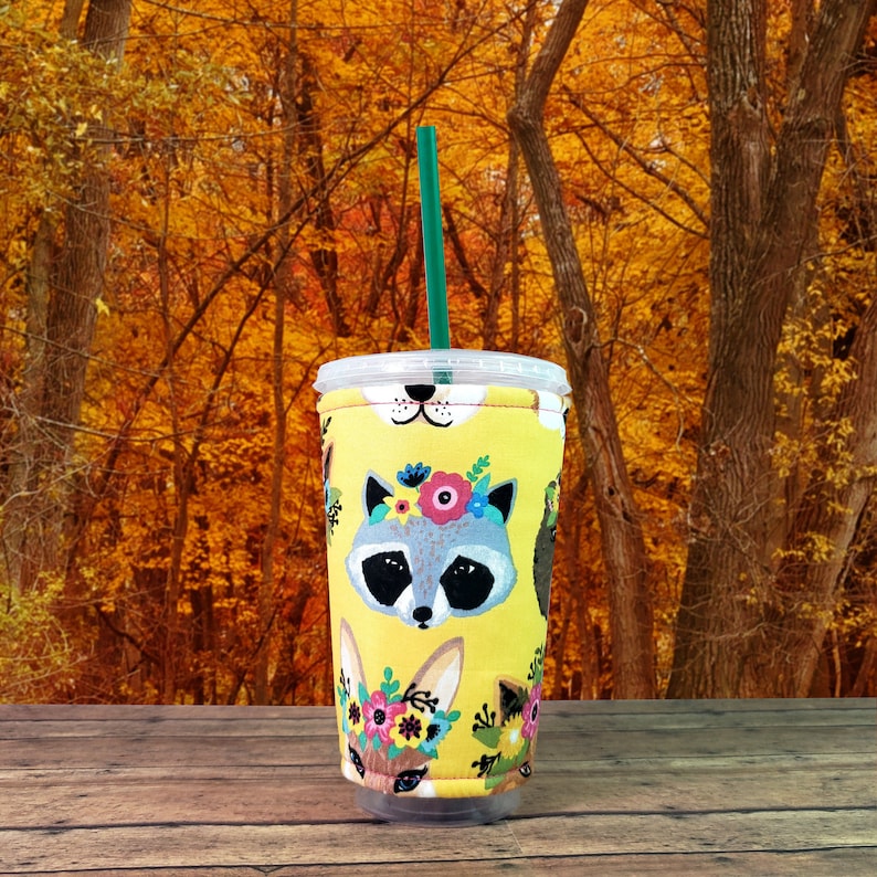 Puede incluir: Un protector de taza reutilizable amarillo con un dise&ntilde;o floral que presenta un mapache, un zorro y un conejo. El protector est&aacute; sobre una mesa de madera con un fondo borroso de follaje oto&ntilde;al.