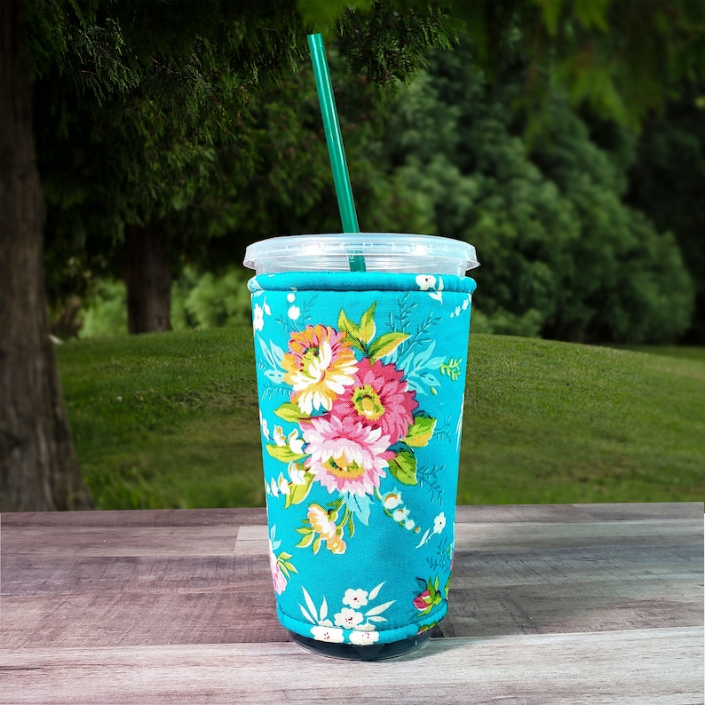 Puede incluir: Un protector de taza reutilizable de color azul turquesa con un estampado floral con flores rosas, blancas y amarillas. El protector est&aacute; hecho de tela y tiene una costura cosida alrededor de la parte superior.