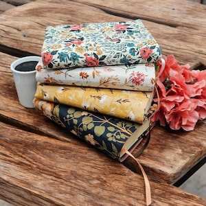 May include: A stack of four rectangular books with fabric covers featuring floral and botanical patterns in shades of cream, yellow, and dark blue, accompanied by a white cup and a cluster of pink flowers with green leaves on a wooden surface.