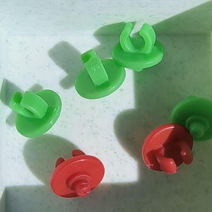 May include: An overhead shot shows five plastic fasteners in a white container. The fasteners are in two colors: green and red. The green fasteners have a circular base with a curved clip, while the red ones have a similar base with a different clip design.