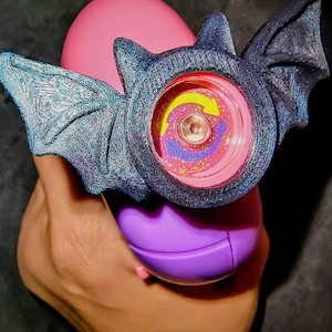 以下が含まれることがあります： コウモリ型のデザインのノベルティおもちゃ。おもちゃは、カラフルなスパイラル模様と黄色の矢印が付いた円形の中心部を持っています。コウモリの羽は、キラキラとしたアクセントが付いた濃い青色です。おもちゃはピンクと紫のオブジェクトの上に保持されています。