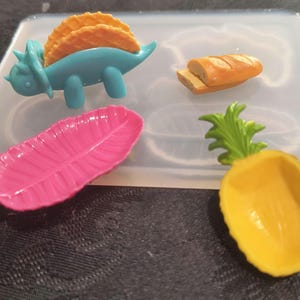 Pode incluir: Um molde de silicone com várias formas temáticas de comida. Inclui um tricerátopo azul segurando tacos, uma pena rosa, um abacaxi amarelo e um pão. O molde é transparente e está sobre uma superfície escura.