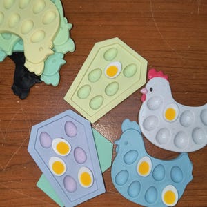 Pode incluir: Cinco brinquedos sensoriais de silicone com formato de frango, caixão e dois estilos diferentes de caixas de ovos. Os brinquedos são todos em cores pastel e têm uma textura com reentrâncias. Cada brinquedo tem um pequeno círculo em cada reentrância, que pode ser empurrado para dentro e para fora.