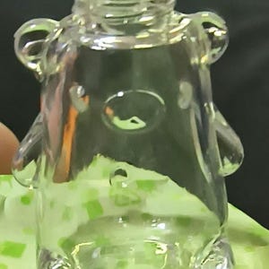 Pode incluir: Uma garrafa transparente em forma de urso de vidro com uma abertura redonda no topo. A garrafa tem orelhas pequenas e arredondadas e uma reentrância circular para o nariz. A garrafa está sobre uma superfície com padrão verde e branco.