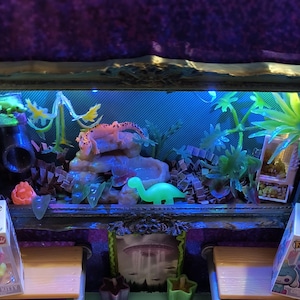 Puede incluir: Un diorama en miniatura con un interior azul brillante, que presenta una figura de dinosaurio verde neón, plantas artificiales y una formación rocosa. Se ven dos cajas de Funko Pop a cada lado.