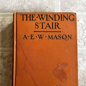May include: The cover of a vintage book titled "The Winding Stair" by A.E.W. Mason. The cover is a dark orange color with a black silhouette of a caravan of camels with riders.