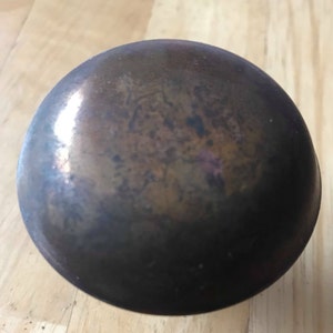 Könnte beinhalten: Eine dunkle, metallische Schüssel mit runder Form. Die Schüssel hat eine gesprenkelte Oberfläche mit Brauntönen, Bronze und einem Hauch von Lila. Die Oberfläche der Schüssel reflektiert das Licht und erzeugt Glanzlichter und Schatten.