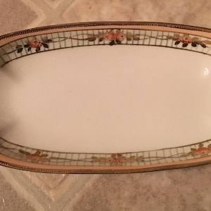 Könnte beinhalten: Eine weiße ovale Servierplatte mit einem goldenen und rosafarbenen Blumendesign. Die Platte hat zwei Griffe an den Seiten.