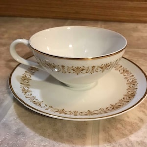 Puede incluir: Un juego de taza y platillo de té blanco con ribete dorado y un delicado patrón floral dorado.