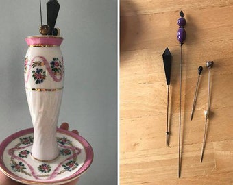 May include: A white porcelain pincushion with a pink rim and floral design. The pincushion is shaped like a vase and has a matching saucer. Four decorative pins with black and purple beads are shown on a wooden surface.