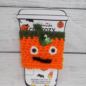 May include: Orange crocheted pumpkin cup cozy with green leaves and black eyes and mouth. The cozy is on a white paper cup with the text "Handmade Cup Cozy" and "Reusable cup cozy to protect hands from hot beverage cups!"