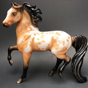 May include: A detailed figurine of a horse in a rearing pose. The horse has a tan coat with white and gold-colored spots. Its mane and tail are black, and the hooves are dark brown. The horse is a collectible item.
