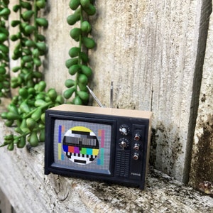 May include: A small, retro-style television set with a black surround and cream-coloured top. The screen shows a multi-coloured test card. The TV has a silver aerial and control dials. The word "Fenyue" is printed on the front.