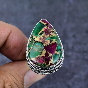 以下が含まれることがあります： 大きな涙型の宝石をあしらったシルバーリング。石は緑、バーガンディ、ゴールドの混合色です。リングは装飾的なシルバーバンドです。鮮やかな色とデザインが印象的なジュエリーです。