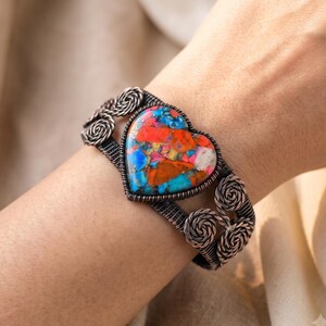 Könnte beinhalten: Ein Armreif mit einem herzförmigen Stein, der ein buntes Mosaikmuster aus leuchtendem Orange, Türkisblau und Weiß aufweist. Das Armband besteht aus dunkel kupferfarbenem Draht, der kunstvoll gewickelt und zu dekorativen Spiralen geformt ist.
