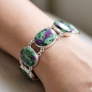 May include: A link bracelet featuring multiple oval-shaped zoisite stones with vibrant magenta and deep green inclusions. Each stone is set in a polished silver-toned metal bezel and connected by small silver-toned metal circular links.