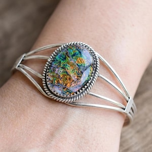 May include: A silver cuff bracelet featuring a central oval-shaped cabochon with a multi-colored iridescent finish in shades of green, blue, and orange. The stone is framed by a twisted rope border and set on a band made of four thin silver wires.