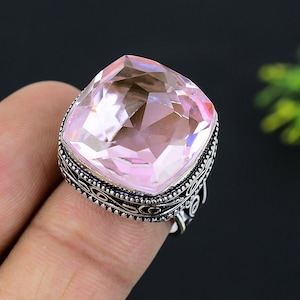May include: A large, square-cut, light pink gemstone ring with a detailed silver band. The gemstone is faceted, catching the light and displaying multiple shades of pink. The ring is set on a finger, showcasing its size and design.