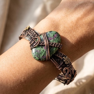 May include: A wire-wrapped cuff bracelet featuring a central oval-shaped green and magenta zoisite stone. The band is constructed from dark copper-toned wire woven into a complex lattice pattern, accented with small brass-colored leaf motifs.