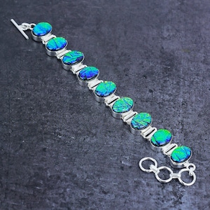 Puede incluir: Pulsera de plata con piedras ovaladas iridiscentes azules y verdes. La pulsera tiene un cierre de palanca y eslabones de conexión. Las piedras están engastadas en un engaste de metal plateado. Mide unos 19 cm.