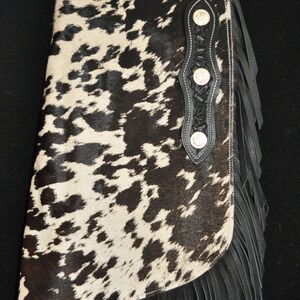 May include: Black and white cowhide chaps with black leather fringe and a black leather belt with a silver buckle. The chaps have a decorative black leather panel with silver studs.