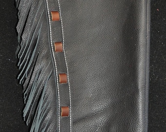 May include: Black leather chaps with decorative brown leather accents and fringe. The chaps have white stitching and brown leather tabs. The top portion features floral and geometric tooling.