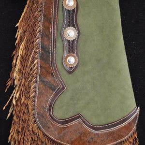May include: Green suede and brown leather western chaps with fringe. The chaps feature decorative silver conchos and a brown leather belt. The chaps have a decorative brown and white stitched border.