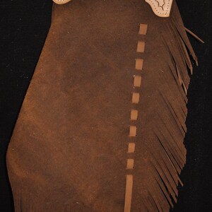 May include: Brown leather chaps with fringe and an oak leaf design on the top flap. The chaps have a belt loop and a buckle.