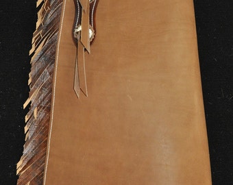 Può includere: Chaps in pelle marrone con dettagli a frangia e un bordo di cucitura bianca. I chaps hanno un design decorativo sulla parte superiore e un passante per la cintura.