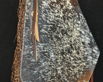 May include: A brown and white cowhide chaps with fringe and a leather strap. The chaps have a brown leather trim and a white stitching detail.