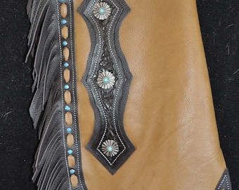 May include: Brown leather chaps with black fringe and decorative stitching. The chaps have a diamond-shaped design with turquoise accents.