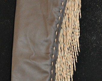 Op de afbeelding: Bruine leren chaps met decoratieve bloemaccenten en franjes. De chaps hebben een bruine leren riem met een zilveren gesp. De franjes zijn lichtbeige van kleur en lopen langs de zijkant van de chaps.
